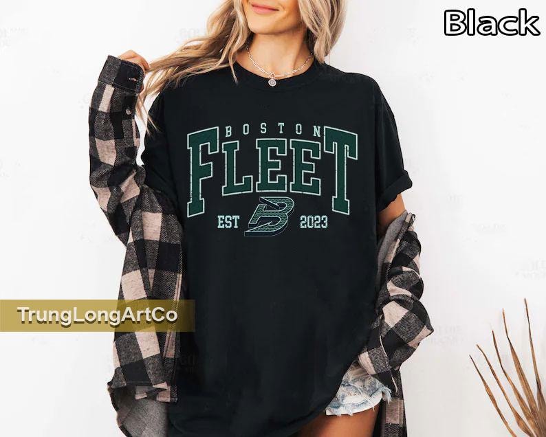 May include: Black t-shirt with the text "BOSTON FLEET" in green and white lettering. The shirt also features the text "EST 2023" and a stylized "B" logo. The shirt is worn with a plaid shirt.