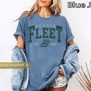 May include: A light blue t-shirt with the text "BOSTON FLEET EST 2023" in green lettering. The shirt has rolled-up sleeves and is worn with a denim jacket. The shirt is part of a clothing collection.