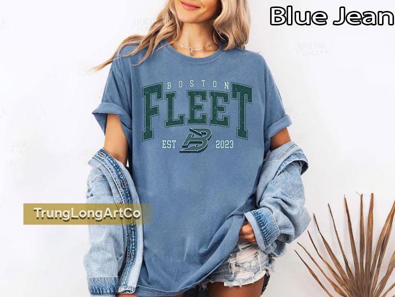 May include: A light blue t-shirt with the text "BOSTON FLEET EST 2023" in green lettering. The shirt has rolled-up sleeves and is worn with a denim jacket. The shirt is part of a clothing collection.