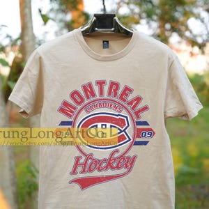 Puede incluir: Una camiseta beige con un logotipo de hockey de los Canadiens de Montreal de estilo vintage. El logotipo presenta el nombre del equipo, "Canadiens", y el n&uacute;mero "09" en un esquema de colores rojo, blanco y azul. Las palabras "Hockey" est&aacute;n escritas en una fuente de letra roja, blanca y azul debajo del logotipo.