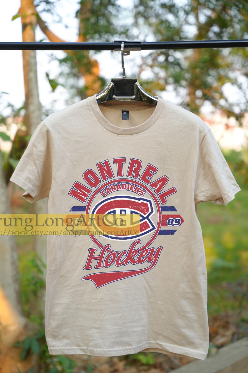 Puede incluir: Una camiseta beige con un logotipo de hockey de los Canadiens de Montreal de estilo vintage. El logotipo presenta el nombre del equipo, "Canadiens", y el n&uacute;mero "09" en un esquema de colores rojo, blanco y azul. Las palabras "Hockey" est&aacute;n escritas en una fuente de letra roja, blanca y azul debajo del logotipo.