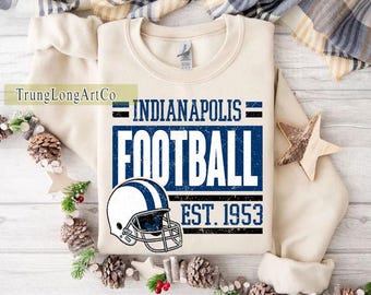 May include: A cream-coloured sweatshirt with the text "INDIANAPOLIS FOOTBALL EST. 1953" in blue and black, and a football helmet graphic. The sweatshirt is decorated with pinecones and a wooden star.