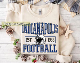 Könnte beinhalten: Cremefarbenes Sweatshirt mit dem blauen Schriftzug "INDIANAPOLIS FOOTBALL", einem Footballhelm-Grafik und dem Text "EST. 1953". Das Sweatshirt wird auf einer weißen Oberfläche mit dekorativen Elementen wie Tannenzapfen und Sternen präsentiert.