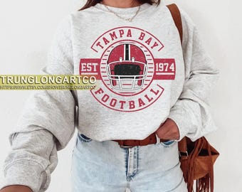 Pode incluir: Camisola cinza claro com um gráfico vermelho e branco de um capacete de futebol e o texto "TAMPA BAY FOOTBALL EST 1974". A camisola tem mangas compridas e gola redonda.