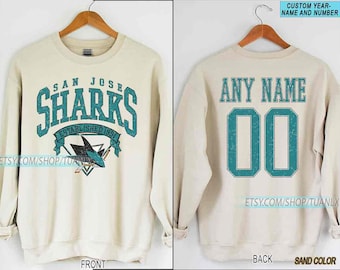 May include: A sand-coloured sweatshirt with the San Jose Sharks logo on the front, and the text "SAN JOSE SHARKS ESTABLISHED 1991". The back shows "ANY NAME" and the number "00" in teal. The sweatshirt is displayed on a wooden hanger.