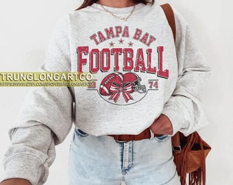 Pode incluir: Um moletom cinza claro com as palavras "TAMPA BAY FOOTBALL" em vermelho, juntamente com um gráfico de futebol americano e capacete. O moletom tem mangas compridas e gola redonda. Os números "74" e "93" também são visíveis.