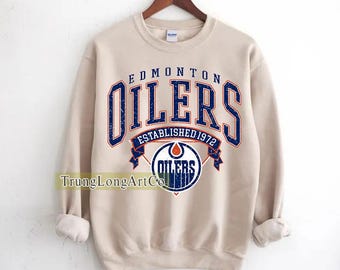 Puede incluir: Sudadera beige con el texto "EDMONTON OILERS ESTABLISHED 1972" en azul y blanco. La sudadera presenta el logotipo de los Oilers en el centro. La marca "GILDAN" es visible en la etiqueta.