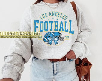 May include: A light gray sweatshirt with the text "LOS ANGELES FOOTBALL" in blue and yellow. The graphic includes a football, helmet, and bow. The sweatshirt has long sleeves and a crew neck.
