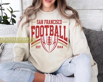 Op de afbeelding: Crèmekleurige sweatshirt met de tekst "SAN FRANCISCO FOOTBALL" in het rood, met een voetbalafbeelding. De sweatshirt heeft ook de tekst "EST 1944" in het rood. De sweatshirt wordt gedragen door een persoon die een blauwe spijkerbroek draagt.