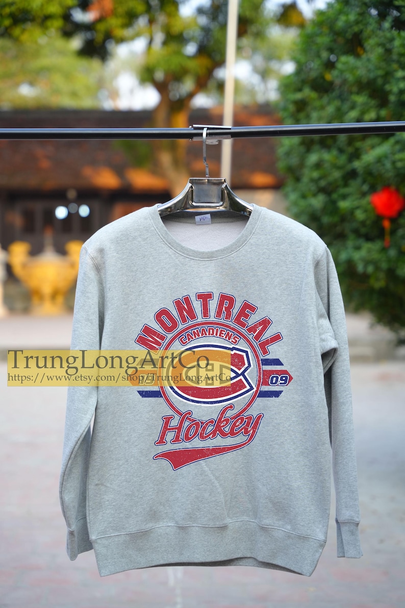 Puede incluir: Una sudadera gris con un logotipo de hockey de los Canadiens de Montreal de estilo vintage. El logotipo presenta el nombre del equipo, "Canadiens", y el n&uacute;mero "09" en rojo, blanco y azul. El texto "Hockey" est&aacute; impreso debajo del logotipo en una fuente roja, blanca y azul.