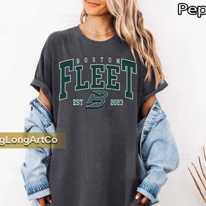 May include: A dark gray t-shirt with the text "BOSTON FLEET" in green block letters. Below the text is "EST 2023" and a green logo. The shirt is worn with a light blue denim jacket.