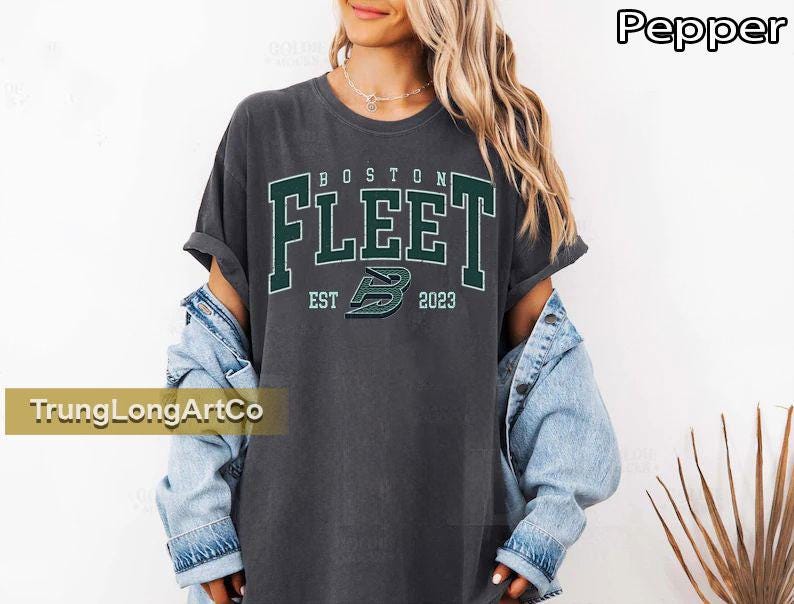 May include: A dark gray t-shirt with the text "BOSTON FLEET" in green block letters. Below the text is "EST 2023" and a green logo. The shirt is worn with a light blue denim jacket.