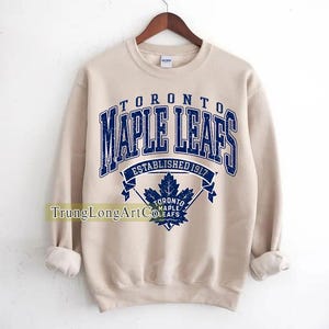 May include: A beige sweatshirt with the text "TORONTO MAPLE LEAFS ESTABLISHED 1917" in blue. Below the text is a blue maple leaf logo. The sweatshirt has long sleeves and a crew neck.