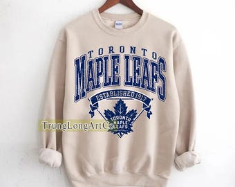 Puede incluir: Sudadera beige con la inscripción azul "TORONTO MAPLE LEAFS ESTABLISHED 1917". Debajo, un logotipo de hoja de arce azul. La sudadera tiene mangas largas y cuello redondo.