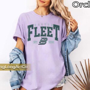 May include: A light purple t-shirt with the text "BOSTON FLEET EST 2023" in a dark green, collegiate-style font. The shirt is worn with a denim jacket. The word "Orchid" is in the top right corner.