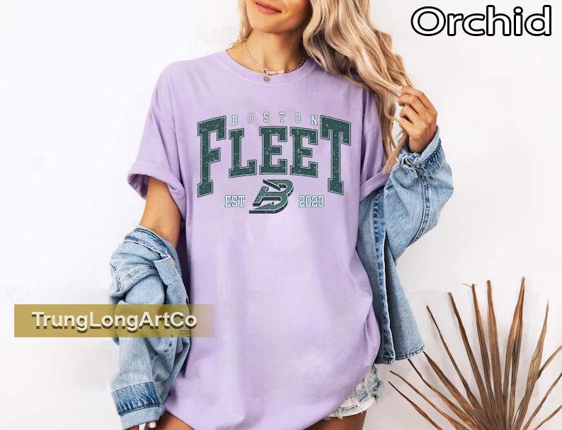May include: A light purple t-shirt with the text "BOSTON FLEET EST 2023" in a dark green, collegiate-style font. The shirt is worn with a denim jacket. The word "Orchid" is in the top right corner.