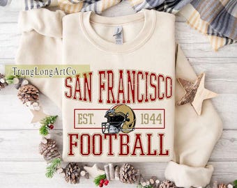 Op de afbeelding: Crèmekleurige sweatshirt met de rode woorden "SAN FRANCISCO FOOTBALL" en een afbeelding van een footballhelm. De tekst "EST. 1944" is ook opgenomen. De sweatshirt is op een wit oppervlak gevouwen, met decoratieve elementen.
