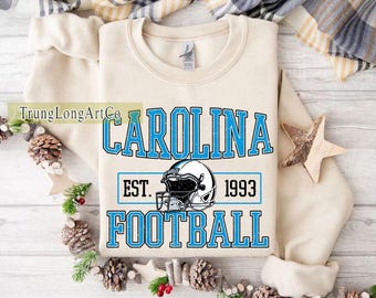 May include: A cream-coloured sweatshirt featuring the text "CAROLINA FOOTBALL EST. 1993" in light blue. A football helmet graphic is centered below the text. The sweatshirt is folded on a white surface with decorative elements.