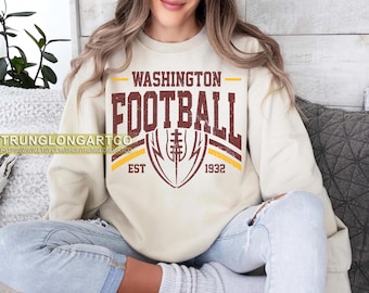 May include: A cream-colored sweatshirt featuring the text "WASHINGTON FOOTBALL" in a distressed maroon font, with a football graphic and the year "1932." The design is in a vintage style. The person is wearing blue jeans.