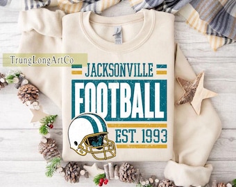 May include: A cream-coloured sweatshirt with the text "JACKSONVILLE FOOTBALL EST. 1993" in teal and gold. A football helmet graphic is below the text. The sweatshirt is decorated with pinecones, stars, and greenery.