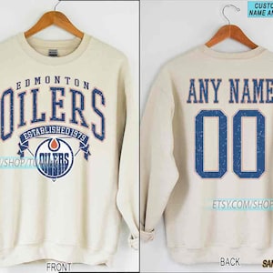 May include: Sand-coloured sweatshirt with the Edmonton Oilers logo on the front, featuring the word "OILERS" in blue. The back shows "ANY NAME" above the blue number "00". The sweatshirt is displayed on a wooden hanger.