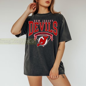 Może przedstawiać: Ciemnoszara koszulka z czerwonym napisem "NEW JERSEY DEVILS", logo i napisem "ESTABLISHED 1974". Koszulka ma wygląd vintage.