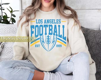 May include: A cream-colored sweatshirt with the text "LOS ANGELES FOOTBALL" in blue and yellow. The design includes a football graphic and the year "1959". The sweatshirt is worn by a person.