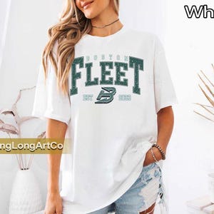 May include: White t-shirt with the text "BOSTON FLEET" in green block letters. The shirt also features the text "EST 2022" and a stylized "B" logo. The shirt is a casual style.