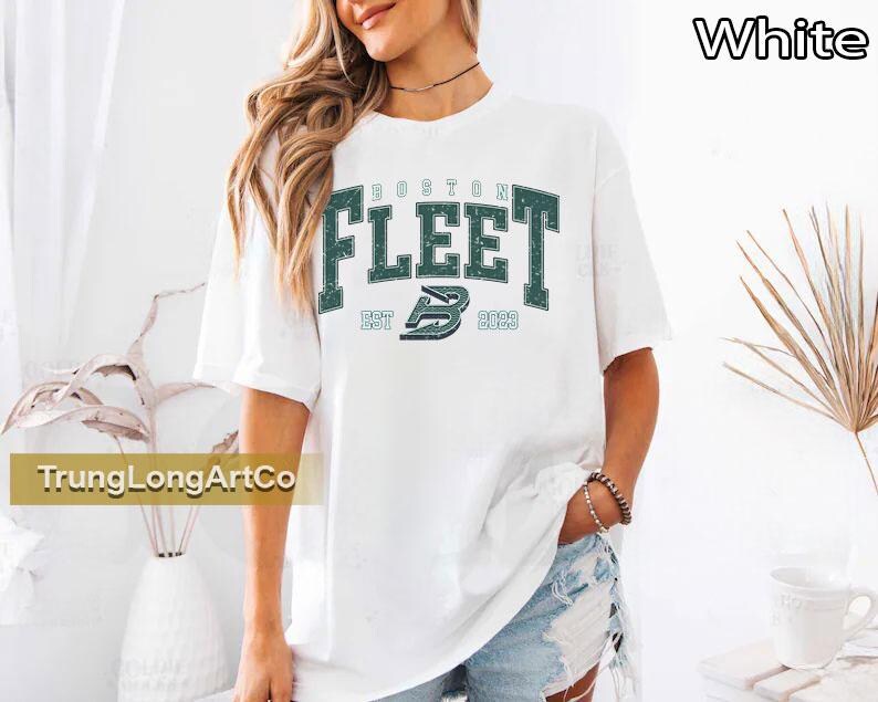 May include: White t-shirt with the text "BOSTON FLEET" in green block letters. The shirt also features the text "EST 2022" and a stylized "B" logo. The shirt is a casual style.