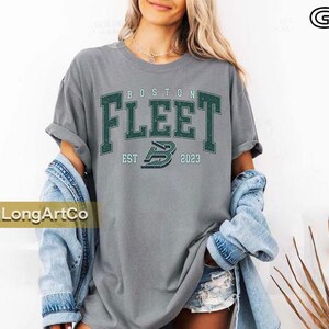 May include: Grey t-shirt with the text "BOSTON FLEET" in green block letters. Below the text is "EST 2023" and a logo. The model is wearing a denim jacket.