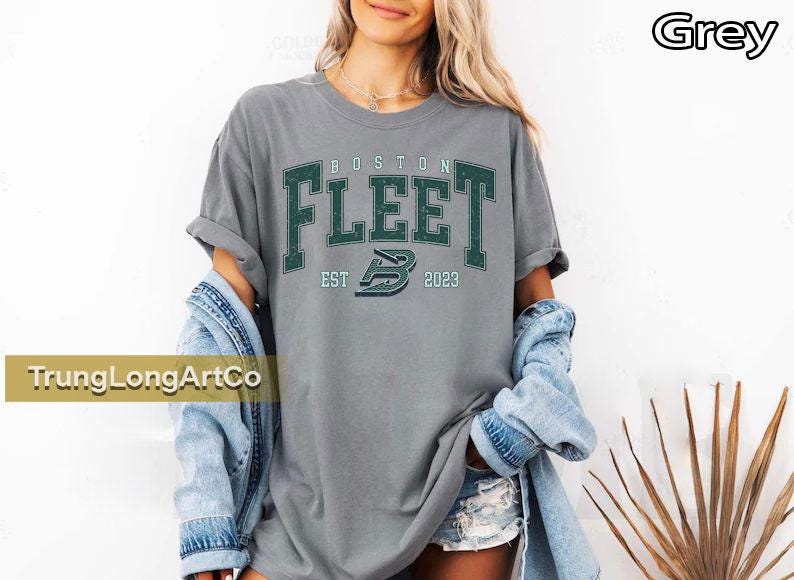 May include: Grey t-shirt with the text "BOSTON FLEET" in green block letters. Below the text is "EST 2023" and a logo. The model is wearing a denim jacket.
