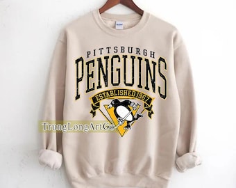 May include: A tan sweatshirt featuring the text "PITTSBURGH PENGUINS" in black and yellow, with a penguin logo. The words "ESTABLISHED 1967" are below the logo. The sweatshirt is hanging on a wooden hanger.