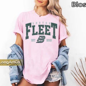 May include: Pink t-shirt with the text "BOSTON FLEET" in dark green, with "EST 2023" below. The shirt is worn with a denim jacket and shorts. The word "Blossom" is in the top right corner.