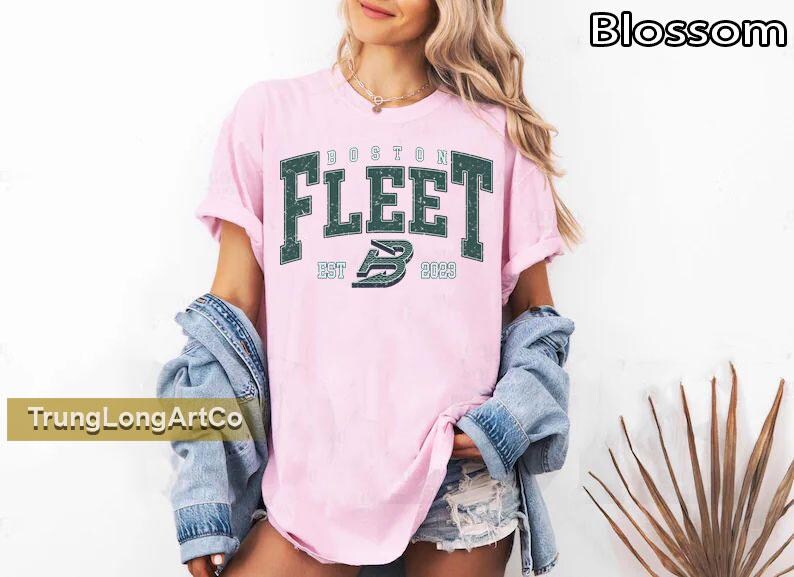 May include: Pink t-shirt with the text "BOSTON FLEET" in dark green, with "EST 2023" below. The shirt is worn with a denim jacket and shorts. The word "Blossom" is in the top right corner.