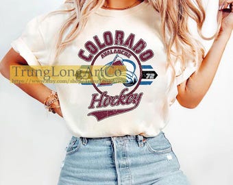 May include: A white t-shirt with a vintage-style graphic of the Colorado Avalanche hockey team logo. The logo features the team's name, "Colorado Avalanche", in a maroon circle with a hockey stick and puck. The text "Hockey" is written in maroon below the circle.