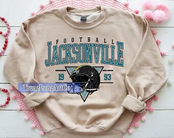May include: A beige sweatshirt featuring the word "FOOTBALL" above "JACKSONVILLE" in teal. A black football helmet graphic is below, with the numbers "19" and "93" on either side. The sweatshirt has a crew neck and long sleeves.