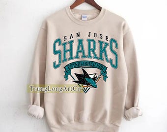 May include: A beige sweatshirt with the text "SAN JOSE SHARKS" in teal lettering. The design includes a shark logo and the words "ESTABLISHED 1991". The sweatshirt is hanging on a wooden hanger.
