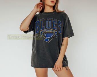 Pode incluir: Camiseta cinza escuro com a inscrição "ST. LOUIS BLUES" em azul e dourado, e o texto "ESTABLISHED 1967". A camiseta também apresenta um logotipo e um visual vintage desgastado.