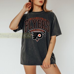 May include: Dark gray t-shirt featuring the text "PHILADELPHIA FLYERS" in red and black, with the team logo. The shirt also includes the text "ESTABLISHED 1967". The shirt has a vintage, worn look.