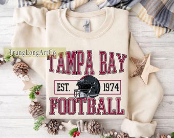 Pode incluir: Camisola creme com o texto "TAMPA BAY FOOTBALL" em bordô, um gráfico de capacete de futebol e o ano de "1974". O design tem um visual vintage. Uma peça de roupa casual e confortável.