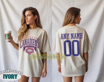 Puede incluir: Camiseta de color marfil con el logotipo de los Canadiens de Montreal en rojo, blanco y azul. La parte trasera de la camiseta muestra el texto "ANY NAME" encima del número "00" en azul. La camiseta es de la marca Comfort Colors.