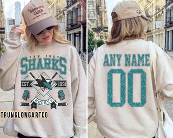 May include: A sand-coloured sweatshirt featuring "SAN JOSE SHARKS" and hockey graphics. The back displays "ANY NAME" above the number "00". The sweatshirt also has the text "EST. 1991" and "HOCKEY".