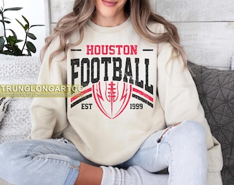 Könnte beinhalten: Cremefarbenes Sweatshirt mit dem Text "HOUSTON FOOTBALL" in Schwarz und Rot sowie einer Football-Grafik. Das Sweatshirt hat auch den Text "EST 1999".