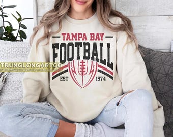 Pode incluir: Camisola creme com o texto "TAMPA BAY FOOTBALL" em fonte preta desgastada, e um gráfico de futebol vermelho e preto. A camisola também inclui o texto "EST 1974".