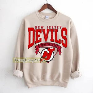 Puede incluir: Sudadera beige con la inscripción "NEW JERSEY DEVILS" en letras rojas. Debajo, una pancarta roja dice "ESTABLISHED 1974". También se ve el logo de los Devils, una "N" roja dentro de un círculo blanco.