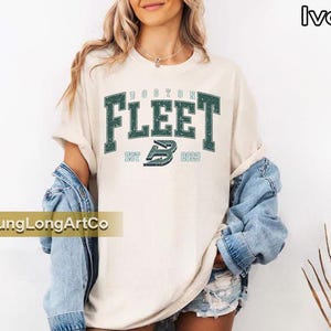 May include: Ivory-colored t-shirt with the text "BOSTON FLEET" in dark green block letters. Below the text is "EST 2023" and a stylized "B" logo. The shirt is worn with a denim jacket.