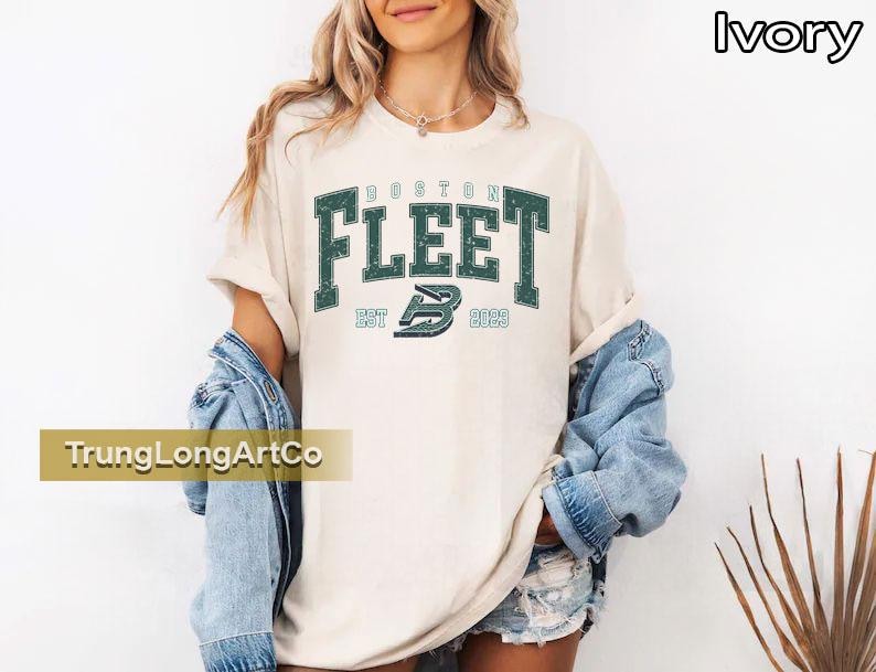 May include: Ivory-colored t-shirt with the text "BOSTON FLEET" in dark green block letters. Below the text is "EST 2023" and a stylized "B" logo. The shirt is worn with a denim jacket.