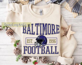 May include: Cream-coloured sweatshirt with the text "BALTIMORE FOOTBALL" in blue, and a football helmet graphic. The text "EST. 1996" is also included. The sweatshirt is a crew neck style.