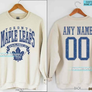 May include: Sand-coloured sweatshirt featuring "TORONTO MAPLE LEAFS" in blue lettering on the front, with a maple leaf emblem and "ESTABLISHED 1917." The back displays "ANY NAME" and the number "00" in blue. The sweatshirt is shown on a wooden hanger.