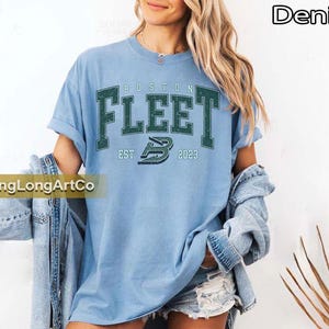 May include: Light blue t-shirt with the text "BOSTON FLEET" in dark green block letters. The shirt also features the text "EST 2023" and a stylized "B" logo. The word "Denim" is in the upper right corner.